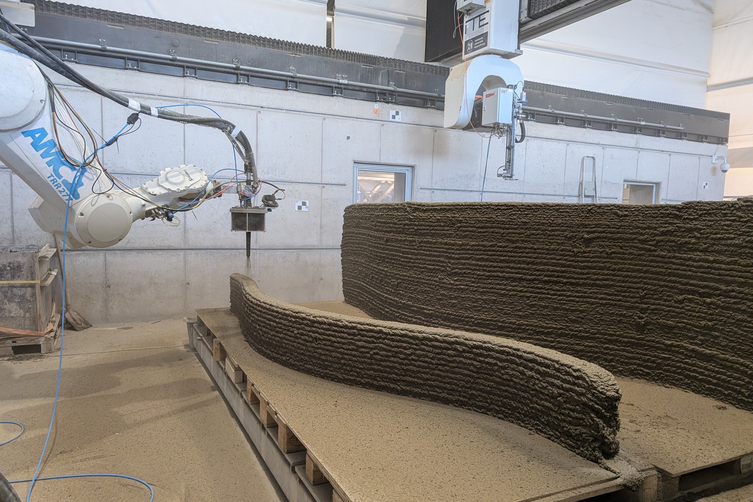 Industrieroboter druckt Beton schichtweise zu gebogener Wand in Werkhalle, 3D-Betondruck im Einsatz.