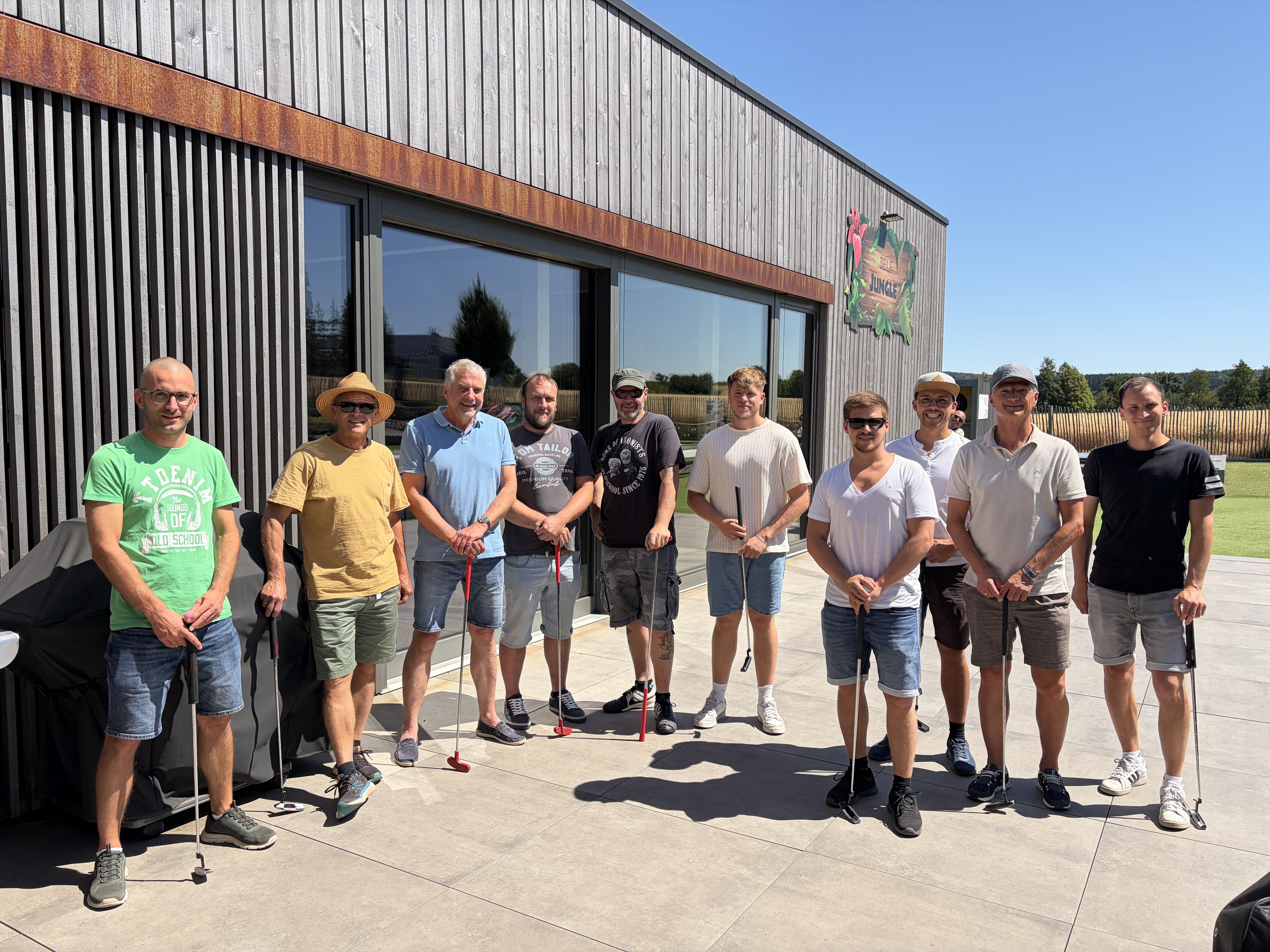 Gruppe von zehn Personen mit Minigolfschlägern vor einem modernen Holzgebäude bei Sonnenschein.