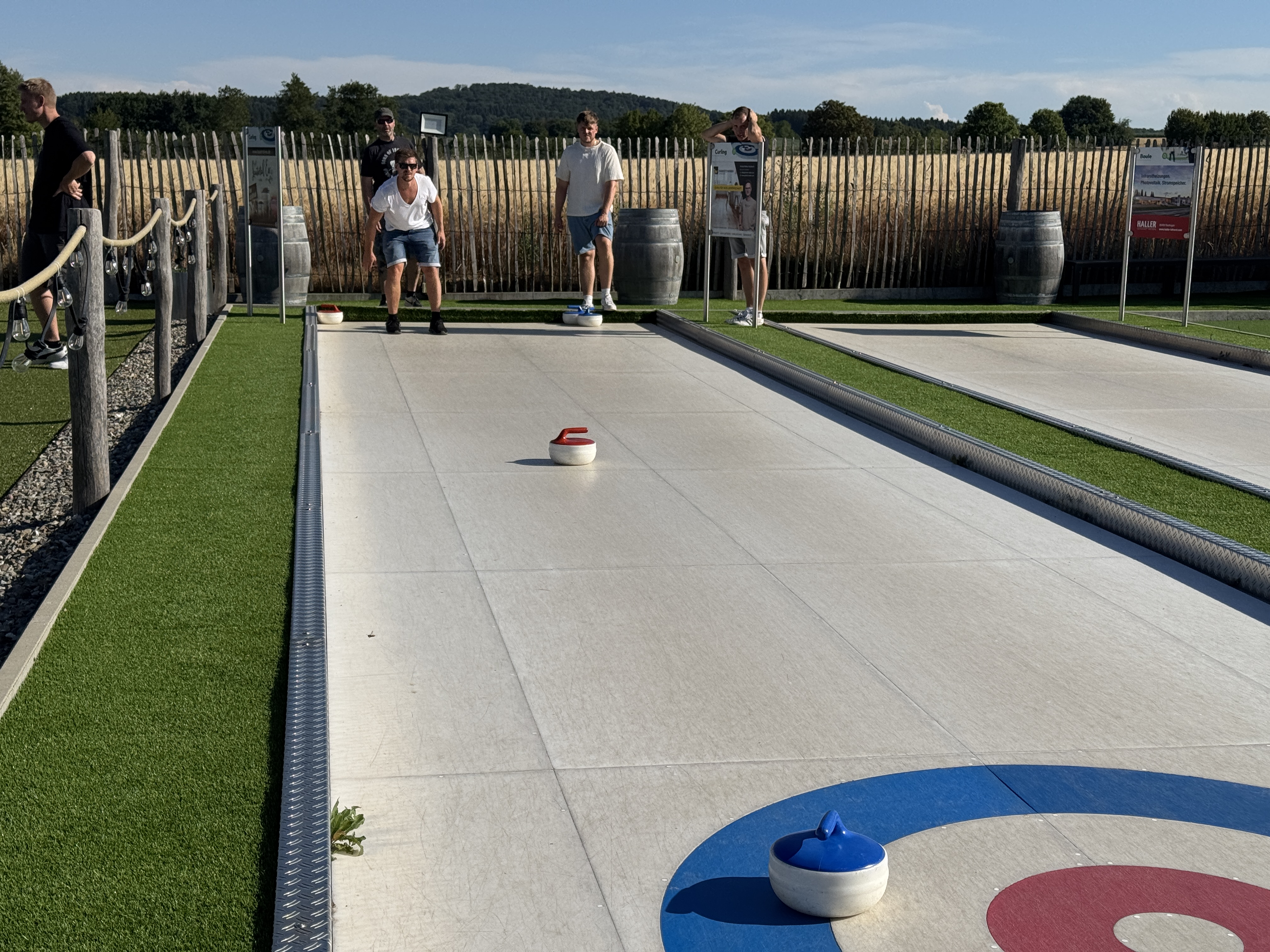 Personen spielen Eisstockschießen auf einer Outdoor-Bahn vor Feldern und Bäumen bei sonnigem Wetter.