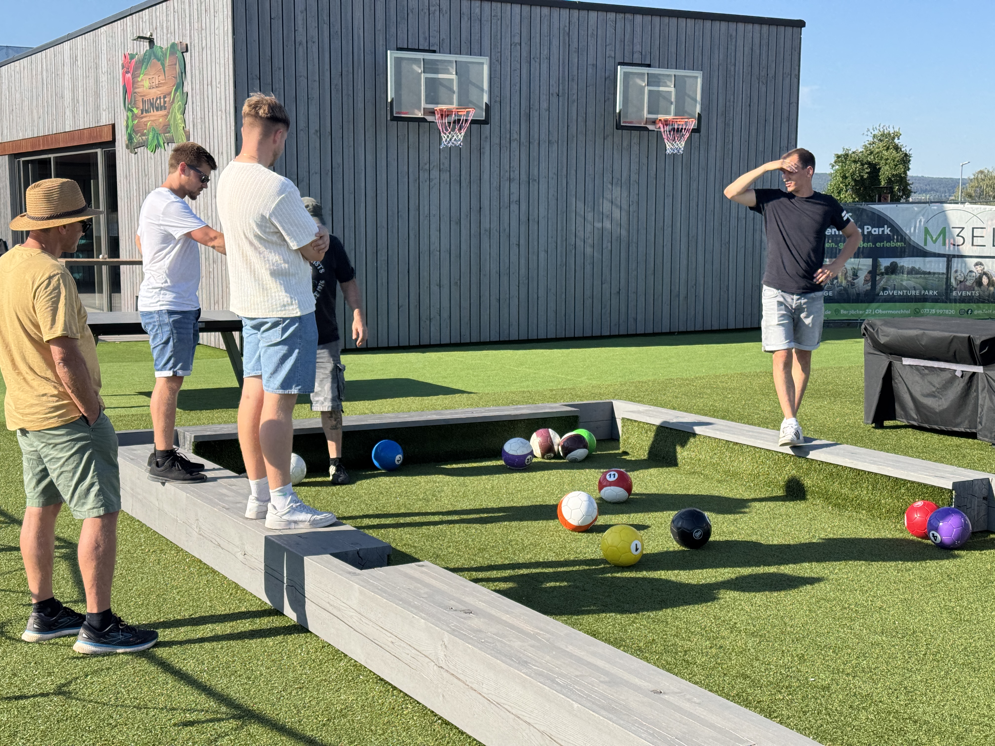 Mehrere Personen spielen Fußball-Billard auf einer Outdoor-Anlage mit großen Bällen und Holzbanden.
