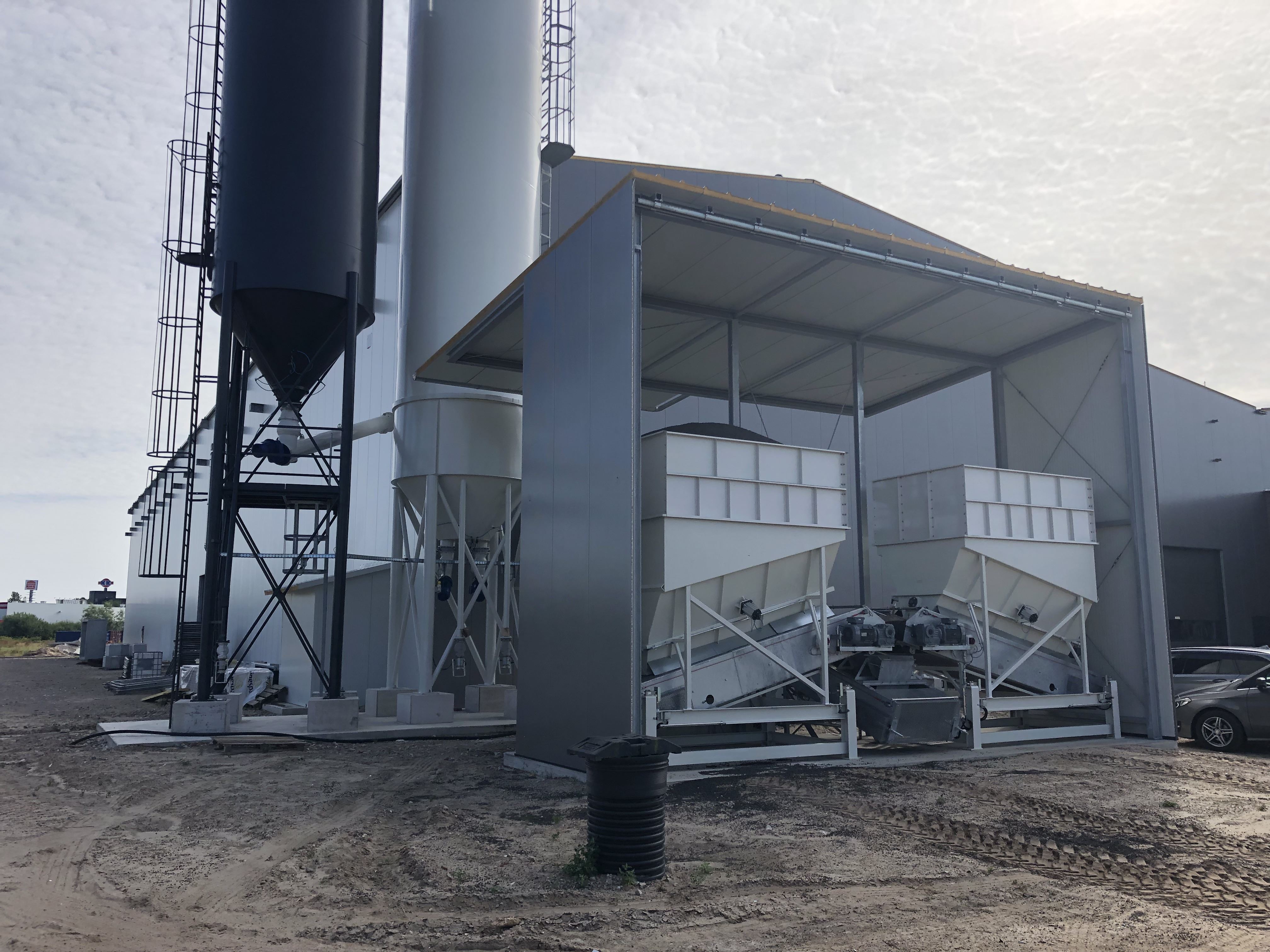 Industrieanlage mit Silos, Dosierbehältern und überdachter Fördertechnik auf einem Betriebsgelände.