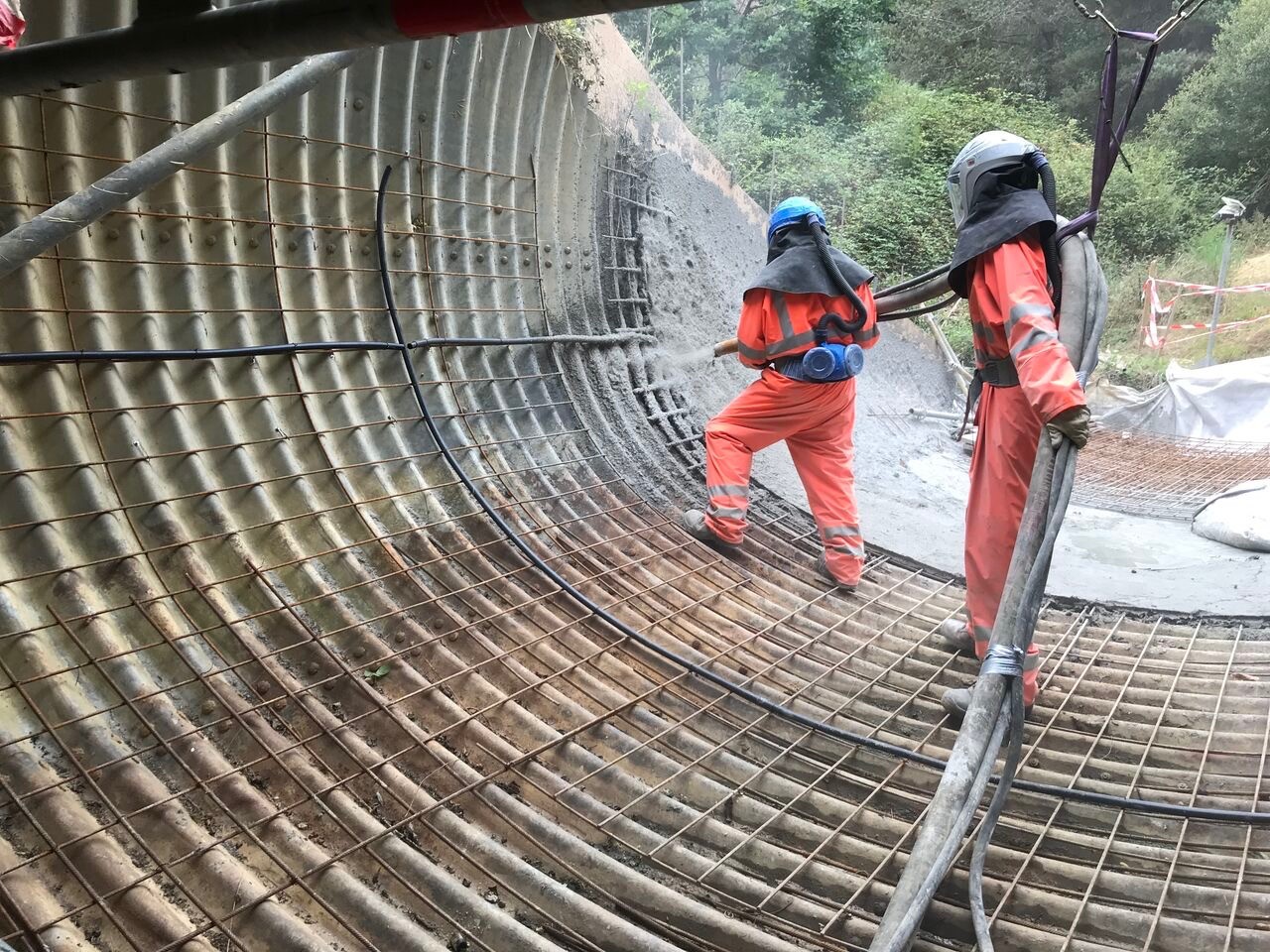 Two skilled workers are applying shotcrete to a tunnel or pipe structure lined with reinforcing steel.