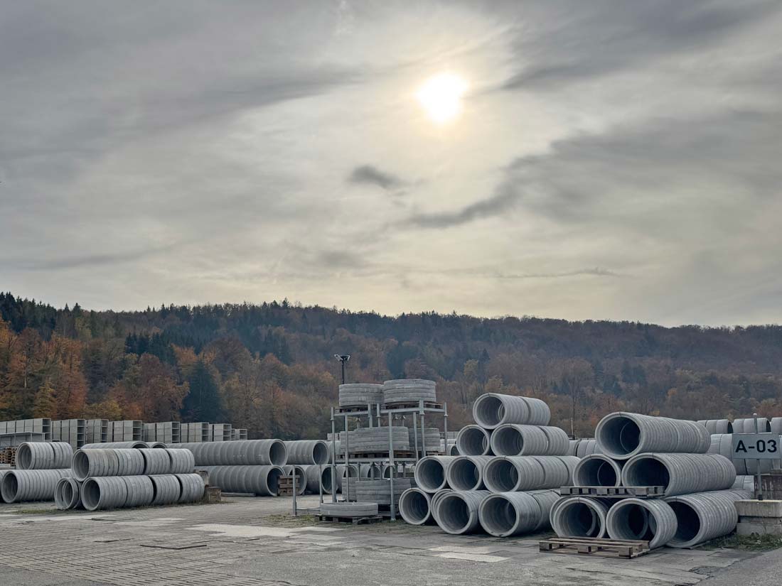 Weißer Roboterarm von Decke mit Spruehduese vorne aus welcher Beton sprüht. Nebelige Umgebung und Paletten auf dem Boden.