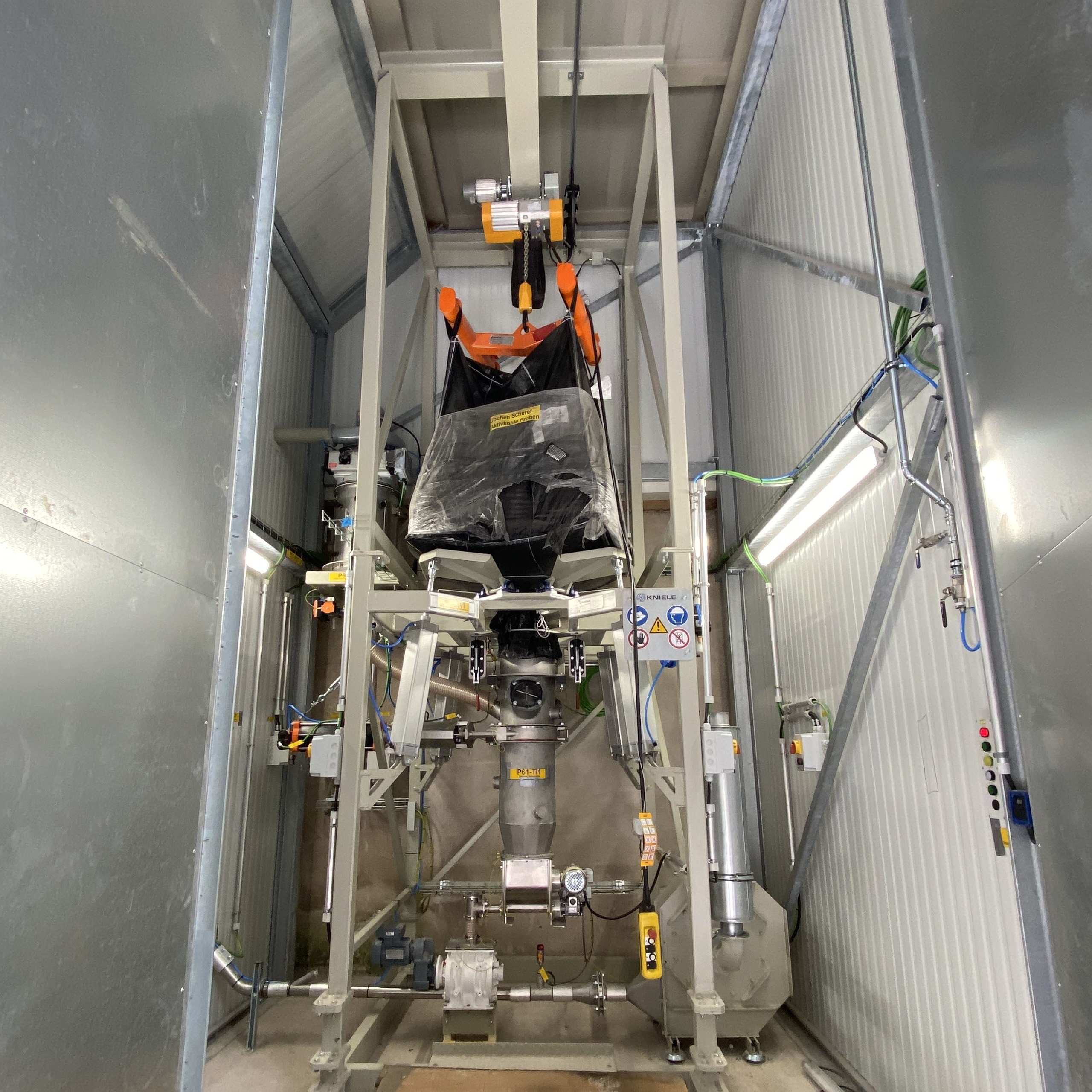 Big-bag station with crane, orange crossbeam, black big bag in a confined enclosure